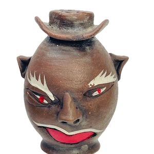 May include: A brown ceramic face-shaped container with a small hat on top. The face has red eyes, a red mouth, and white accents around the eyes. The container has ear-like protrusions on the sides.