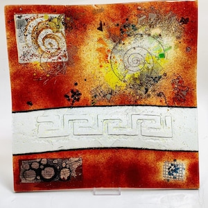 May include: A square glass plate with a red, orange, and yellow background. The plate has a white border with a Greek key design. The plate has a variety of abstract designs in white, black, and green.