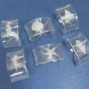 May include: Six clear acrylic napkin rings with etched sea life designs, including a seahorse, seashell, starfish, and other marine creatures. The napkin rings are displayed on a blue surface.