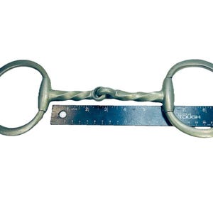 May include: A silver-colored horse bit with a twisted center and large circular rings on each end. The bit is approximately 9 inches long, as measured by a ruler placed beneath it. The bit is designed for use in equestrian activities.