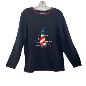 May include: Black long-sleeve sweater with a lighthouse design. The lighthouse is red and white with a green top and blue flag. The sweater has red trim around the neckline and cuffs. The design includes waves and seagulls.