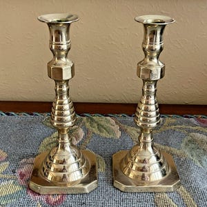 May include: Two silver-colored candlesticks. Each candlestick has a square base, a ribbed, conical midsection, and a hexagonal element. The top has a flared, circular cup to hold a candle. The candlesticks are displayed on a floral patterned surface.