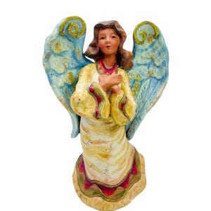 May include: A ceramic angel figurine with light blue wings and gold glitter accents. The angel wears a yellow robe with red and green trim and holds its hands together. The figurine has brown hair and a serene expression.