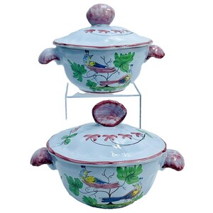 May include: Two white ceramic bowls with lids, each featuring a hand-painted design of birds on a vine with green leaves. The lids have a pink knob and a pink rim.