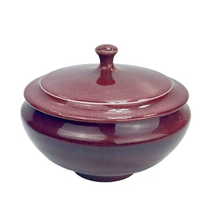 Puede incluir: Un cuenco de cerámica rojo oscuro con tapa. El cuenco tiene un acabado brillante y una forma ligeramente redondeada. La tapa tiene un pequeño pomo en la parte superior.