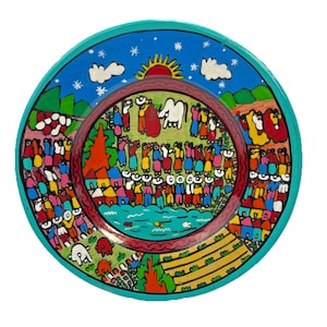 May include: A hand-painted ceramic plate with a vibrant, colorful design. The plate features a central scene of people, a white animal, and a body of water, surrounded by a turquoise border. The artwork includes a sun, clouds, and stars.