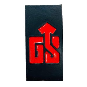 May include: A rectangular black object with a glossy red logo. The logo features the letters "GS" with an upward-pointing arrow. The object has a textured surface and is set against a white background.