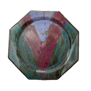 May include: An octagonal ceramic plate with a colorful glaze. The plate features a central design of purple and blue, surrounded by a teal and blue border. The plate has a glossy finish and is likely handmade.
