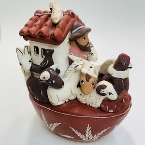 May include: A ceramic figurine of a man with a red roofed house on his back, carrying a variety of animals including a sheep, a goat, a horse, a bird, and a dog. The figurine is painted in brown, white, and black.