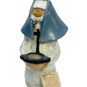 May include: A ceramic figurine of a nun, featuring a blue veil and a white habit. The figurine holds a small, dark-rimmed bowl. A tag with text about "Poppets" is attached. The figurine has a smiling face.