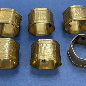 May include: Six brass napkin rings with a hammered texture and a hexagonal shape are arranged on a blue surface. One ring is a different color, possibly silver. The rings are for table decor.