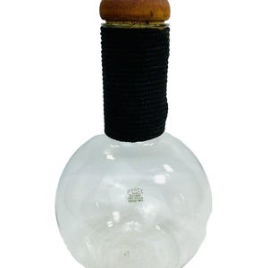 May include: A clear glass bottle with a round body and a cork stopper. The bottle is wrapped with black rope and has a wooden top. The bottle is labeled "PYREX GLASSWARE MADE IN U.S.A.  SERIAL NO. 7131".
