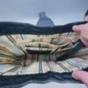 May include: The interior of a black handbag featuring a beige lining with a vertical striped pattern in shades of brown, blue, and white. The bag includes a central zippered compartment and open side pockets for organization within the main space.