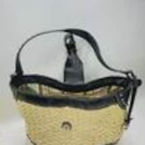 May include: A hobo-style handbag featuring a woven straw body in a light beige tone, accented with black leather trim, a black leather shoulder strap, and a central metallic emblem. The bag has a curved top opening and reinforced black leather corners.