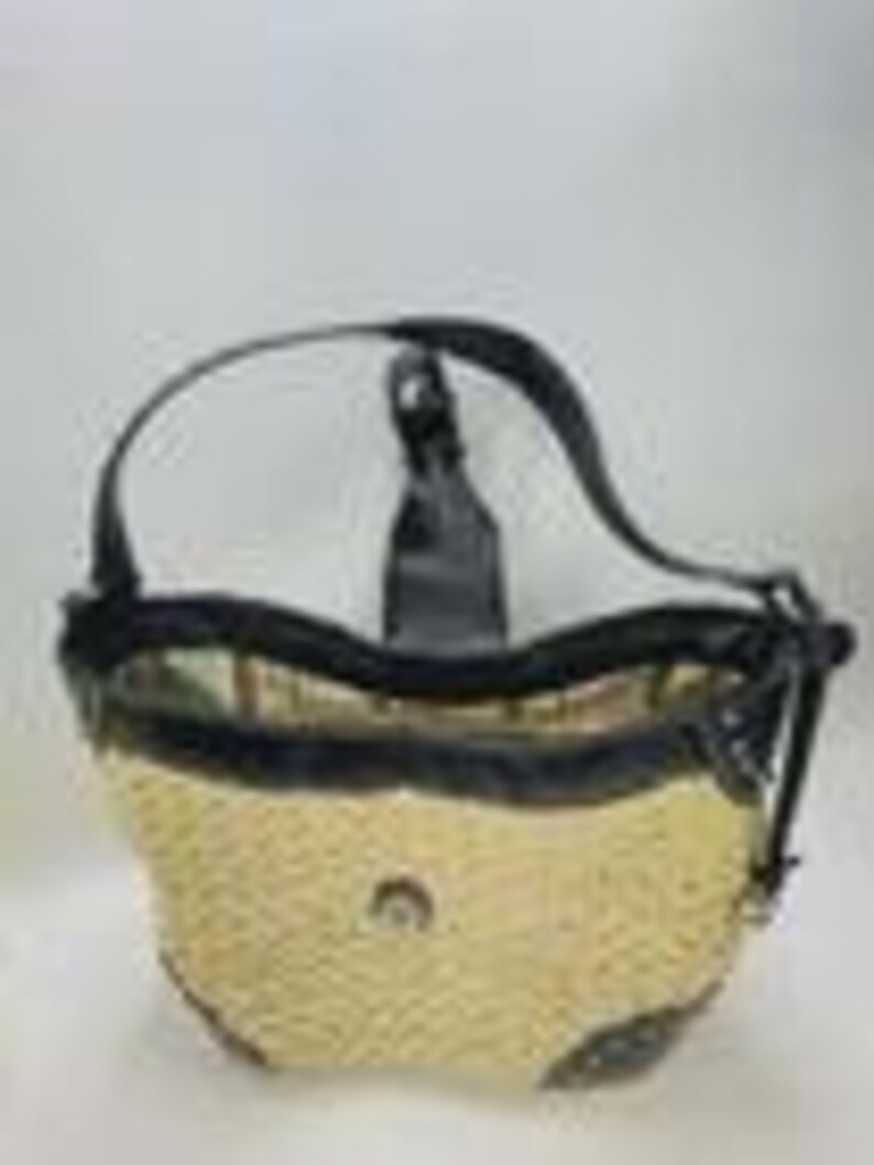 May include: A hobo-style handbag featuring a woven straw body in a light beige tone, accented with black leather trim, a black leather shoulder strap, and a central metallic emblem. The bag has a curved top opening and reinforced black leather corners.