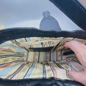 May include: A black leather handbag with a wide strap and a magnetic snap closure. The interior is lined with a cream-colored fabric featuring vertical stripes in light blue, pink, and brown, along with a repeating black swirl pattern.