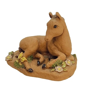 May include: A brown ceramic figurine of a horse foal lying down on a bed of green grass with yellow flowers. The foal has a brown mane and tail and is looking to the left.