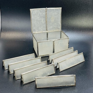 May include: A set of clear glass name card holders and a matching box with a hinged lid. The box and name card holders have a silver metal trim. The name card holders are rectangular and stand upright.
