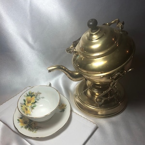 May include: A gold-toned brass teapot with a wooden knob on the lid and a decorative chain. The teapot is sitting on a white surface with a white teacup and saucer with a floral design.
