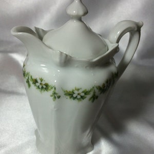 Könnte beinhalten: Ein weißer Porzellan-Milchkännchen mit Deckel, verziert mit einem grün-weißen Blumendesign. Der Milchkännchen hat einen geriffelten Boden und einen zarten Griff.