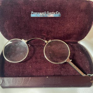May include: Open maroon velvet eyeglasses case with a pair of round, silver-rimmed spectacles. The case has a silver nameplate that reads "Pinkham & Smith Co. Boston". The glasses are open, resting inside the case.