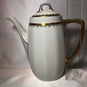 May include: White porcelain coffee pot with gold trim and a black and gold floral design. The pot has a lid and a handle. The pot is 9 inches tall.