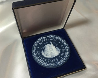 May include: A blue and white ceramic medallion with a white image of a building and the text "METTLACH 1980 COLLECTORS SOCIETY" in a circle.