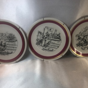 May include: Three white ceramic plates with a red and gold rim. Each plate features a black and white illustration of a musical scene. The first plate shows a group of musicians playing instruments. The second plate shows a group of people singing. The third plate shows a group of people dancing.