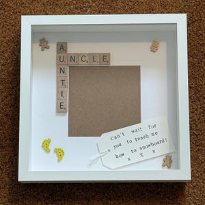 以下が含まれることがあります： 「AUNTIE」と「UNCLE」の文字が書かれたスクラブルタイルの白いフレームのシャドーボックス。フレームには写真用の空白スペースと装飾的な木製の形が含まれています。タグには「Can't wait for you to teach me to snowboard!」と書かれています。