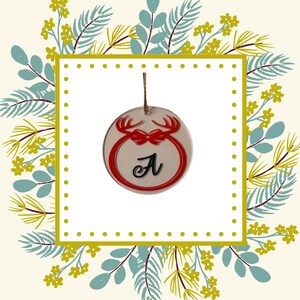 Peut inclure: Une décoration de Noël ronde et transparente avec un motif de renne rouge et un nœud rouge. La lettre "A" est en écriture noire. L'ornement est suspendu à une ficelle marron. Le fond est un motif floral avec un cadre carré jaune.