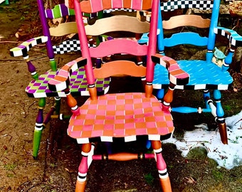 May include: A collection of colorful, hand-painted wooden chairs. Each chair features a unique design, including checkered patterns, stripes, and vibrant color combinations like pink, orange, blue, green, and purple. The chairs have decorative finials and armrests.