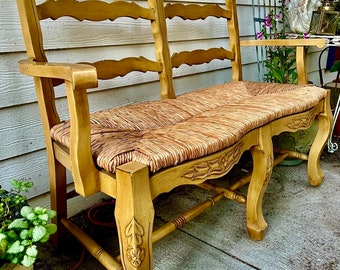 May include: A wooden bench with a woven straw seat and carved details. The bench is painted in a light brown color and has a curved back.