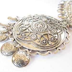Puede incluir: Collar de plata con un gran colgante adornado y pequeños abalorios en forma de moneda. El colgante presenta un diseño detallado e intrincado.
