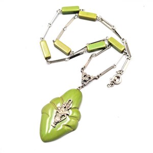Puede incluir: Un collar vintage con un colgante verde claro y un diseño de jarrón plateado. El collar tiene una cadena plateada con eslabones rectangulares verdes y plateados. El colgante es una pieza única y llamativa.