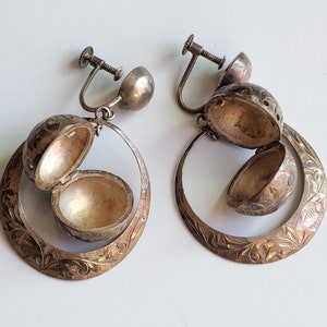 以下が含まれることがあります： フィリグリーのデザインが施された銀製のイヤリングのペア。各イヤリングには、丸いヒンジ付きのロケットが付いており、開くと小さな丸いコンパートメントが現れます。