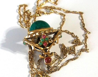Puede incluir: Un collar de cadena de oro con un colgante que presenta una gran piedra preciosa verde rodeada de piedras preciosas rojas y verdes más pequeñas.