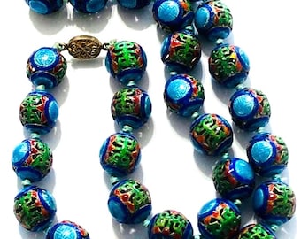 Puede incluir: Un collar largo hecho de cuentas redondas de esmalte azul, verde y rojo con intrincados patrones. Las cuentas están enhebradas en un cordón delgado y tienen un cierre dorado.