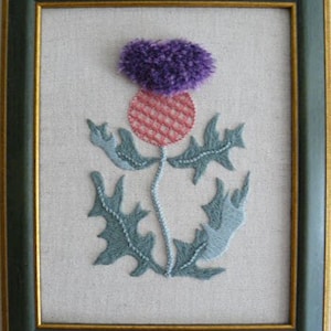 May include: A framed embroidery piece featuring a thistle design on a light beige fabric background. The thistle has a textured purple top, a pink cross-stitched base, and grey-green leaves and stem, all enclosed in a dark green frame.