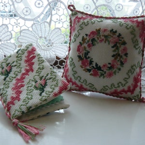 May include: Two small, square, white needlepoint pillows with pink and green floral embroidery. One pillow features a wreath of flowers, the other has a repeating pattern of flowers and leaves.