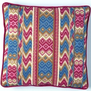 Op de afbeelding: Vierkant decoratief kussen met een levendig, geometrisch patroon. Het ontwerp heeft verticale strepen in blauw, roze, beige en crème, met een effen roze rand. Het kussen is waarschijnlijk gemaakt van geweven stof.