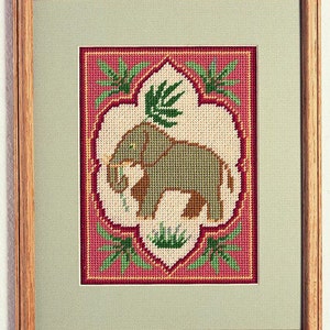 May include: A cross-stitch embroidery design featuring a gray elephant in a pink and green frame. The elephant is standing on a patch of green grass and is surrounded by green leaves.