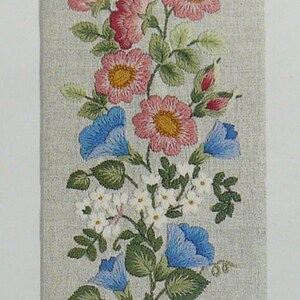Pode incluir: Tapeçaria de parede floral bordada com flores rosa, azuis e brancas e folhas verdes sobre um fundo bege. As bordas superior e inferior têm acabamentos decorativos em metal. Uma peça decorativa para casa.