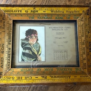 May include: A vintage wooden frame with a yellow and brown ruler border. The frame contains a calendar from February 1924 for Wilmington Fibre Specialty Co. in Wilmington, Delaware. The calendar features a black and white illustration of a woman with a floral headband.