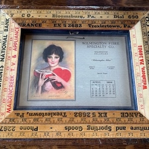 May include: A vintage wooden frame with a calendar from April 1924 for Wilmington Fibre Specialty Co. The calendar features a painting of a woman holding a heart-shaped box of chocolates. The frame is decorated with yellow and white rulers with measurements in inches.