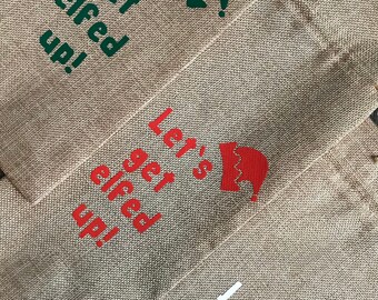 May include: Three burlap gift bags with the text "Let's get elfed up!" in green, red, and white. Each bag has a different colored elf hat graphic.