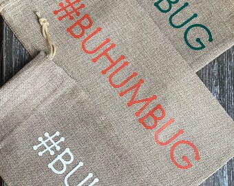 May include: Three burlap gift bags with the hashtag #BUHUMBUG written on them in different colors. The top bag has green text, the middle bag has orange text, and the bottom bag has white text.