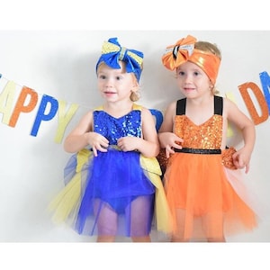 May include: Two young children wearing matching dresses with sequined bodices and tulle skirts. One dress is blue and yellow, the other is orange. Both children wear matching headbands with large bows. A banner with the word "HAPPY" is visible.
