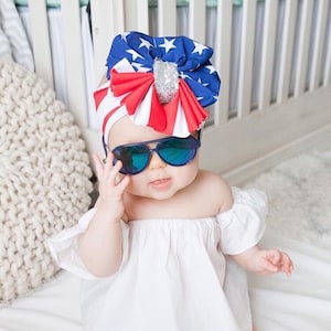 Peut inclure: Un bébé portant des lunettes de soleil aviateur bleues et un bandeau patriotique avec un nœud représentant le drapeau américain. Le bandeau est rouge, blanc et bleu.