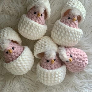 May include: Five crocheted pink chicks in white eggshell-shaped containers. The chicks have black eyes, yellow beaks, and white fluffy tufts on their heads. The eggshells are textured and have a fluffy appearance.