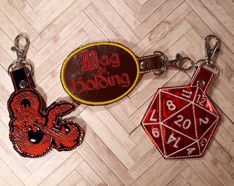 May include: Three keychains featuring a red and black Dungeons and Dragons dice, a red and black D&D character, and a brown leather keychain with the text "Bag of Holding" in red embroidery.